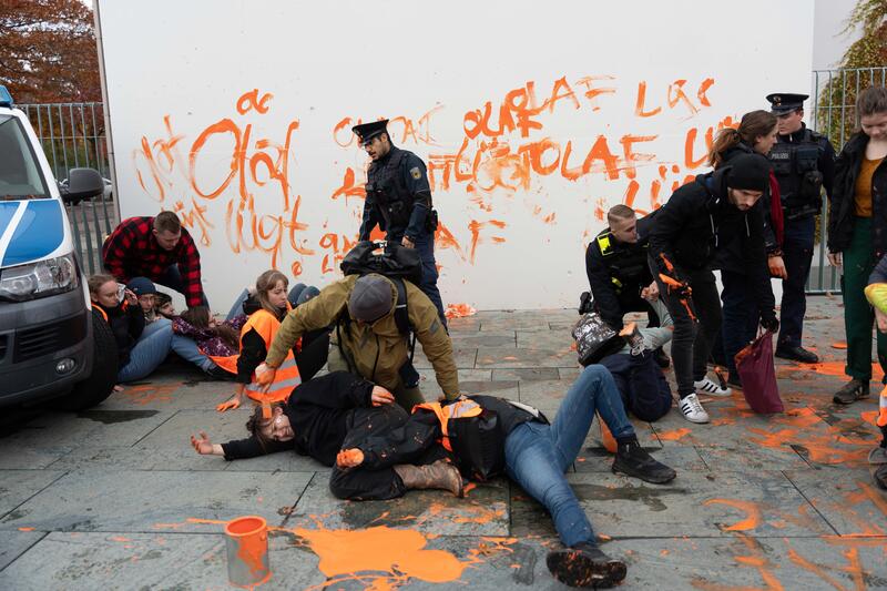 Zivipolizist geht gewaltsam gegen Aktivistinnen vor und schmiert mit dem Pinsel ins Gesicht einer Aktivistin. Über 70 weiblich gelesene Personen der Letzten Generation haben am Dienstag, den 31.10.2023 mit oranger Farbe die Worte “Olaf lügt” viele Male auf die weiße Fassade sowie auf den Boden vor dem Bundeskanzleramt in Berlin gepinselt. Viele von ihnen standen vor den Schriftzügen und halten Plakate in die Höhe. Mit ihrem Protest machen sie auf die fehlende Klimaschutzpolitik des selbst ernannten "Klimakanzlers" Olaf Scholz aufmerksam.   --   Over 70 female-identified individuals from the Last Generation painted the words "Olaf is lying" in orange on the white facade and the ground in front of the Federal Chancellery in Berlin on Tuesday, October 31, 2023. Many of them stood in front of the writings, holding up posters. With their protest, they draw attention to the lack of climate protection policies by the self-proclaimed "Climate Chancellor" Olaf Scholz.