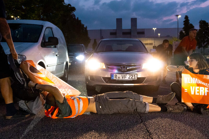 Aktivist*innen der Letzten Generation blockieren am 22.8.2023 in Regensburg die Zufahrten zum BMW-Werk. Sie prangern damit die " ungebremste Verbennung fossiler Brennstoffe " im Verkehrssektor an. In den letzten 30 Jahren habe es dort keine Verbesserungen gegeben. Die LG veranstaltet die Protestphase in Bayern, weil sie hier die größten Blockierer von Klimapolitik sehen. Außerdem fordert ein Tempolimit von 100 km/h auf Autobahnen, die Einführung eines Neun-Euro-Tickets und einen Gesellschaftsrat Klima.   --   Activists of the Last Generation blocked the roads to the BMW factory in Regensburg, Germany. They denounce the " unchecked burning of fossil fuels " in the transport sector. In the last 30 years, they say, there weren't any improvements. The LastGen sees the Bavarian policy makers as largest climate policy blockers. Further the Last Generation demands a speed limit of 100 km/h on highways, the introduction of a nine-euro ticket and a Climate Society Council.