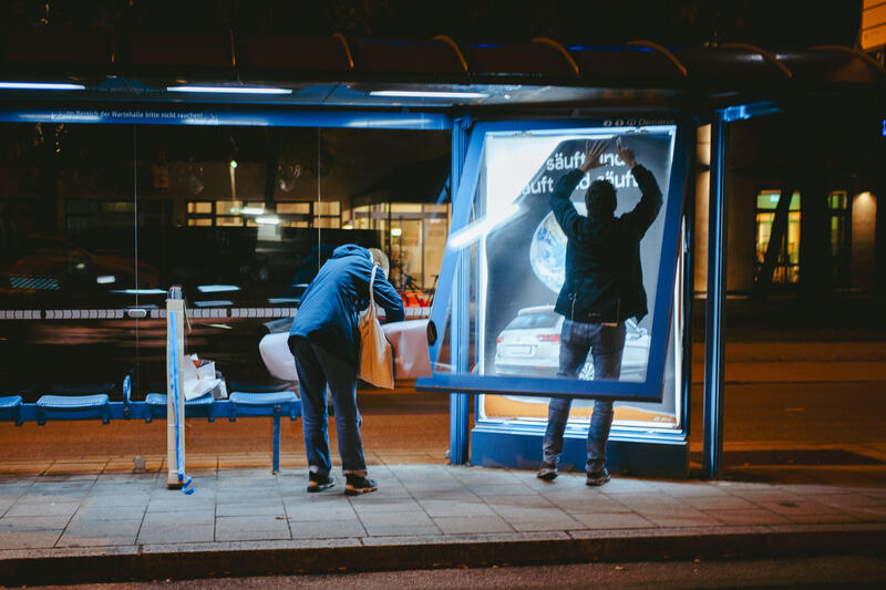 In der Nacht auf den 10.09.2025 versuchten zwei Aktivist:innen von Attac eine Adbusting-Aktion. Sie entfernten einige Werbeplakate und wollten sie durch Plakate gegen die IAA ersetzen. Da sie unvorbereitet waren, konnten sie einige Werbekästen nicht öffnen, einige konnten sie hingegen öffnen, aber ihre Plakate nicht aufhängen. Der Protest von Attac gegen die IAA begleiten sie mit der Froderung, nach einer sozial-ökologischen Wende in der Automobilindustrie und im gesamten Wirtschaftssystem. Sie fordern: Weniger Autos, und stattdessen mehr Platz für Fahrräder und Fußwege. -- On the night of September 10, 2025, two Attac activists attempted an adbusting campaign. They removed several advertising posters and wanted to replace them with posters against the IAA. Since they were unprepared, they were unable to open some of the advertising boxes, while others they were able to open but could not hang their posters. Attac's protest against the IAA is accompanied by demands for a socio-ecological transformation of the automotive industry and the entire economic system. They are calling for fewer cars and more space for bicycles and footpaths instead.