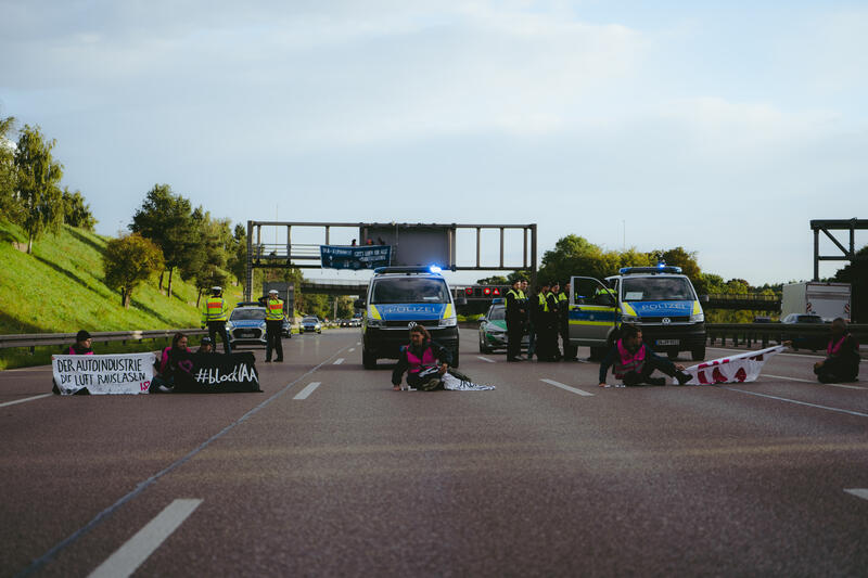 Am 11. September 2025, kurz nach 8, steigen drei Aktivist*innen auf eine Schilderbrücke über der Autobahn nahe der Allianz-Arena in München, Deutschland.
Dazu setzen sich wenig später 6 weitere Aktivist*innen auf die mittlerweile gesperrte Autobahn und kleben sich teilweise fest.
Auch zwei der Aktivist*innen auf der Brücke klebten sich fest, dies verzögerte die Räumung um einiges. Die Aktivist*innen gehörten dem Widerstandskollektiv an und protestierten mit dieser Aktion gegen die IAA, die zurzeit in München stattfindet.  -- On September 11, 2025, shortly after 8 a.m., three activists climb onto a sign bridge above the highway near the Allianz Arena in Munich, Germany.
A little later, six more activists sit down on the now closed highway and partially glue themselves to the ground.
Two of the activists on the bridge also glued themselves to the bridge, which delayed the evacuation considerably. The activists belonged to the resistance collective and were protesting against the IAA, which is currently taking place in Munich.