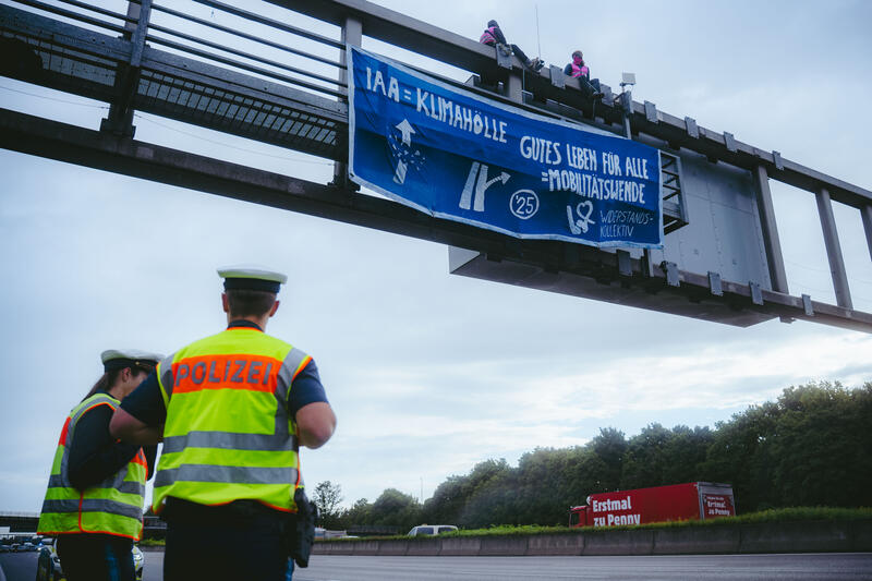 Am 11. September 2025, kurz nach 8, steigen drei Aktivist*innen auf eine Schilderbrücke über der Autobahn nahe der Allianz-Arena in München, Deutschland.
Dazu setzen sich wenig später 6 weitere Aktivist*innen auf die mittlerweile gesperrte Autobahn und kleben sich teilweise fest.
Auch zwei der Aktivist*innen auf der Brücke klebten sich fest, dies verzögerte die Räumung um einiges. Die Aktivist*innen gehörten dem Widerstandskollektiv an und protestierten mit dieser Aktion gegen die IAA, die zurzeit in München stattfindet.  -- On September 11, 2025, shortly after 8 a.m., three activists climb onto a sign bridge above the highway near the Allianz Arena in Munich, Germany.
A little later, six more activists sit down on the now closed highway and partially glue themselves to the ground.
Two of the activists on the bridge also glued themselves to the bridge, which delayed the evacuation considerably. The activists belonged to the resistance collective and were protesting against the IAA, which is currently taking place in Munich.