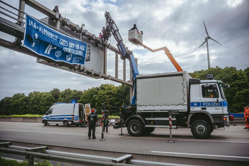 Am 11. September 2025, kurz nach 8, steigen drei Aktivist*innen auf eine Schilderbrücke über der Autobahn nahe der Allianz-Arena in München, Deutschland.
Dazu setzen sich wenig später 6 weitere Aktivist*innen auf die mittlerweile gesperrte Autobahn und kleben sich teilweise fest.
Auch zwei der Aktivist*innen auf der Brücke klebten sich fest, dies verzögerte die Räumung um einiges. Die Aktivist*innen gehörten dem Widerstandskollektiv an und protestierten mit dieser Aktion gegen die IAA, die zurzeit in München stattfindet.  -- On September 11, 2025, shortly after 8 a.m., three activists climb onto a sign bridge above the highway near the Allianz Arena in Munich, Germany.
A little later, six more activists sit down on the now closed highway and partially glue themselves to the ground.
Two of the activists on the bridge also glued themselves to the bridge, which delayed the evacuation considerably. The activists belonged to the resistance collective and were protesting against the IAA, which is currently taking place in Munich.