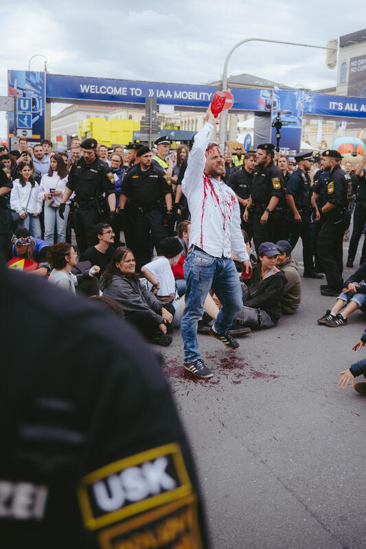 Am 11. September 2025 beteiligten sich um die zwei Dutzend Aktivist*innen an einer Die-In-Aktion von Extinction Rebellion gegen die IAA in München, Deutschland. Dabei legte sich der Großteil auf die Straße und spielte tot. Drei der Aktivist*innen begossen sich mit Mehl, Cola und einem roten Saft, um Staub, Öl und Blut darzustellen. -- On September 11, 2025, around two dozen activists took part in a die-in protest organized by Extinction Rebellion against the IAA in Munich, Germany. Most of them lay down on the street and pretended to be dead. Three of the activists covered themselves in flour, cola, and red juice to represent dust, oil, and blood.