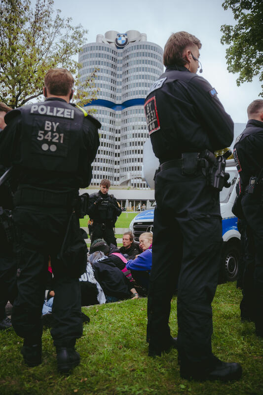 Am 12. September 2025 versammelten sich um die 50 Teilnehmer*innen zu einem Straßenfest gegen die IAA auf einem Grünstreifen vor der BMW-Welt in München, Deutschland. Während der Versammlung kam es immer wieder zu ungehorsamen Aktionen wie dem Besetzen der Straße und dem Klettern auf ein Polizeiauto.  -- On September 12, 2025, around 50 participants gathered for a street party against the IAA on a grass verge in front of BMW Welt in Munich, Germany. During the gathering, there were repeated acts of civil disobedience, such as occupying the street and climbing on a police car.