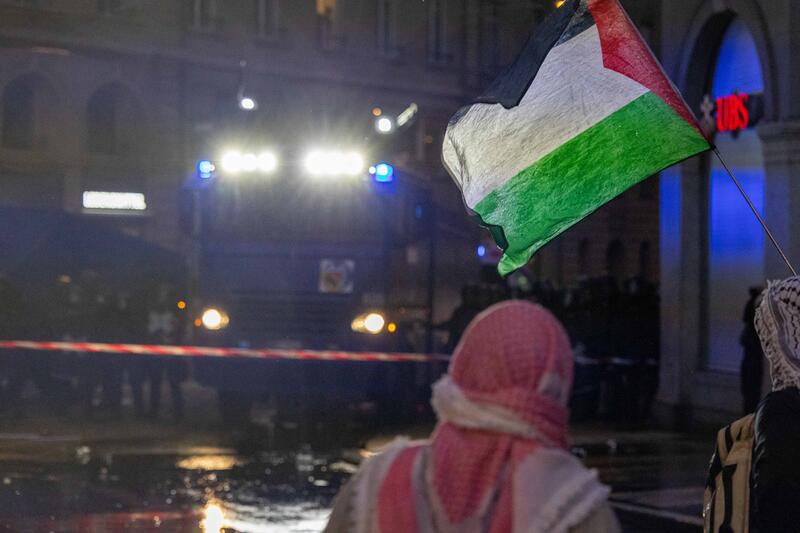 Eine Frau mit Kufiya und eine Palästinaflagge vor dem Wasserwerfer an der Ecke Schauplatzgasse / Bahnhofsplatz. Fotografiert am 11. Oktober 2025.   A woman with a kufiya and a Palestinian flag in front of the water cannon on the corner of Schauplatzgasse and Bahnhofsplatz. Photographed on 11 October 2025.