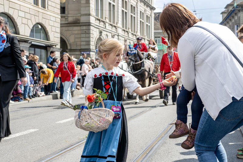 Beim Kinderumzug werden Süssigkeiten und Blumen verteilt, von den Kindern an die Zuschauer, aber auch umgekehrt. Ein Mädchen in Trachtenkleid überreicht einem Kind ein Bonbon. Fotografiert am 19. April 2026.     During the children’s parade, sweets and flowers are handed out – by the children to the spectators, but also the other way round. A girl in traditional dress hands a sweet to a child. Photographed on 19 April 2026.