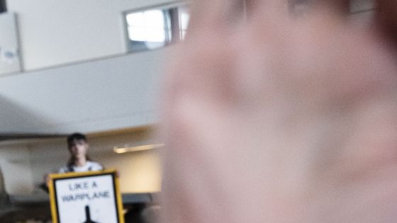 Mann greift in die Kamera des Journalisten während im Hintergrund eine mit Kunstöl-beschmierte Aktivin ein Schild mit der Aufschrift Like A Warplane Oil Kills hochhält.