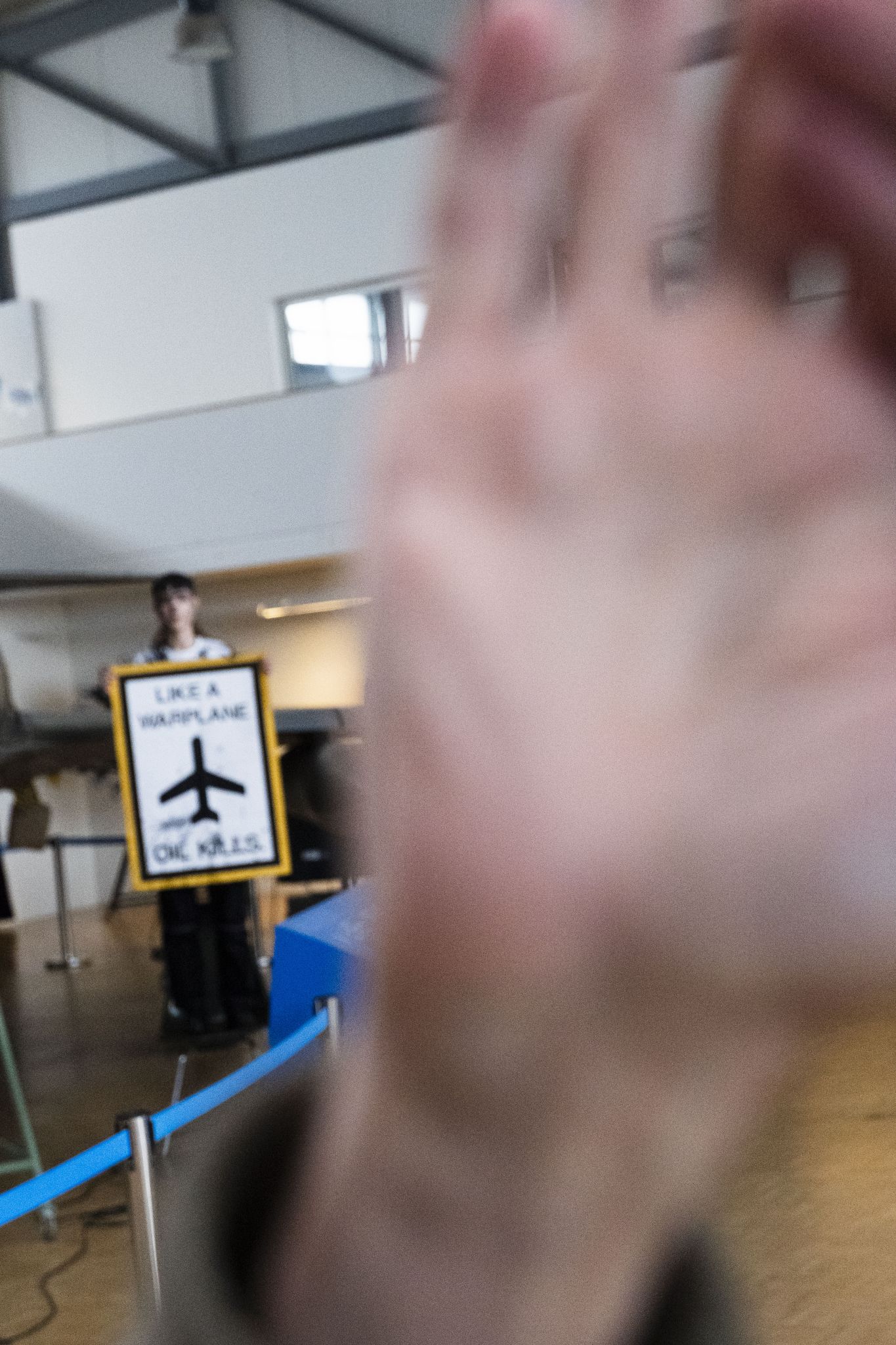 Mann greift in die Kamera des Journalisten während im Hintergrund eine mit Kunstöl-beschmierte Aktivin ein Schild mit der Aufschrift Like A Warplane Oil Kills hochhält.