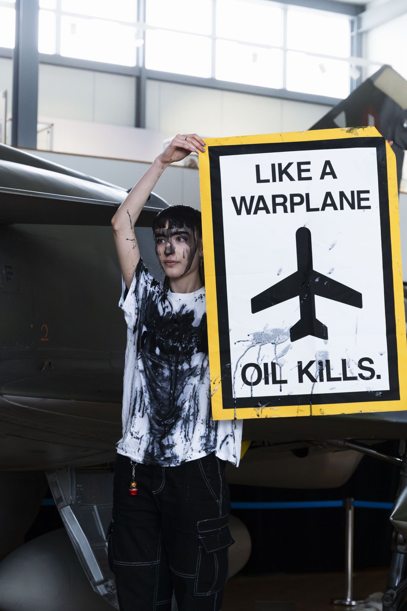 Mit Kunstöl-beschmierte Aktivistin hält Schild mit der Aufschrift Like A Warplane Oil Kills hoch.