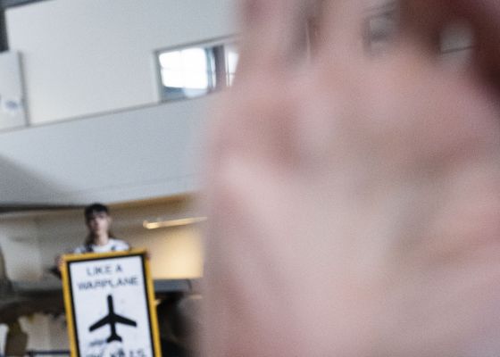 Mann greift in die Kamera des Journalisten während im Hintergrund eine mit Kunstöl-beschmierte Aktivin ein Schild mit der Aufschrift Like A Warplane Oil Kills hochhält.